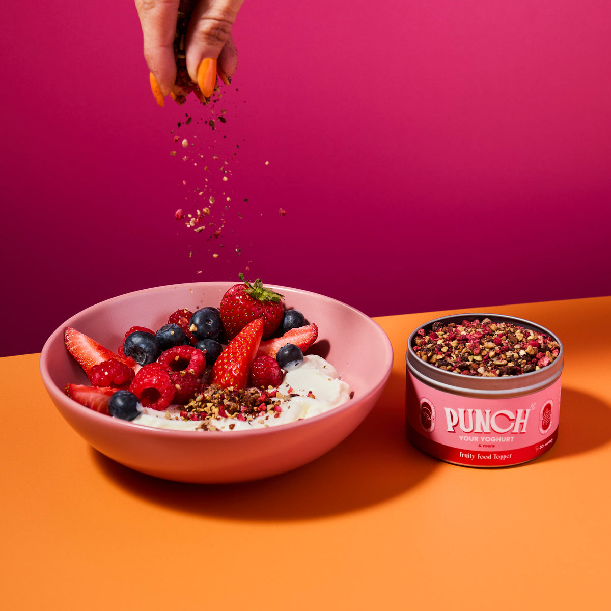 Bowl of fruit salad with yogurt, a tin of Punch your yoghurt fruity food topper, and a hand sprinkling seasoning on a pink and orange background.