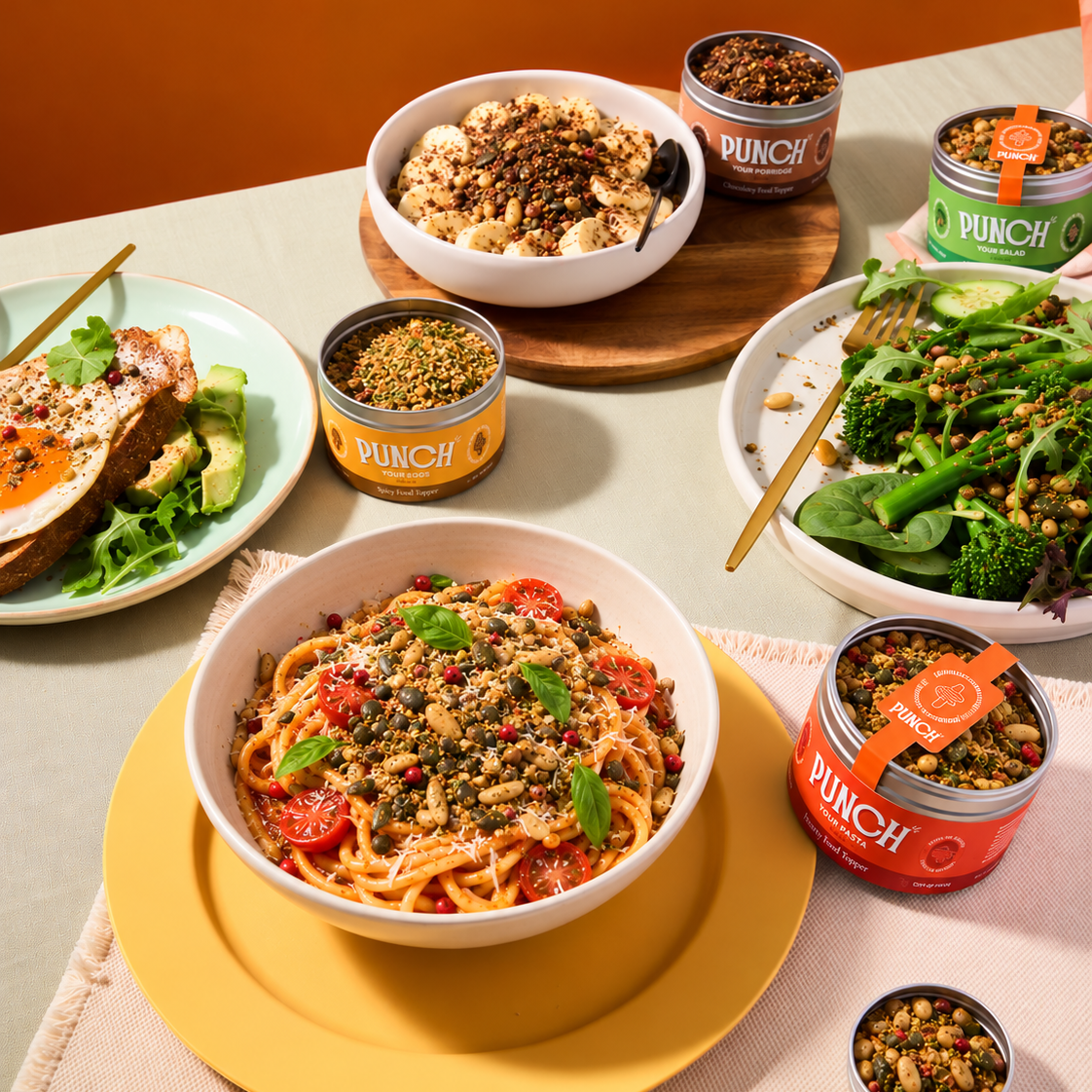 A styled tabletop food scene showing bowls and cans of PUNCH protein powder mixed into pasta, salad, and a breakfast toast. The PUNCH branding is clearly visible on the cans, set against a warm orange wall and beige table setting.