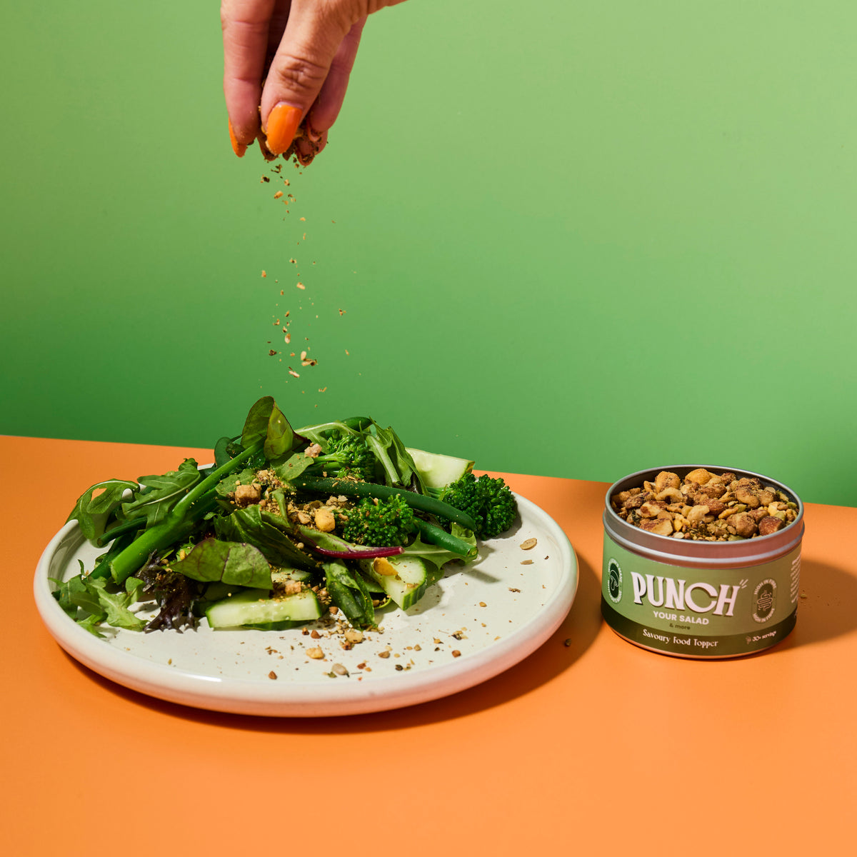 Person adding savoury food topper from a 'Punch your salad' container to a salad on an orange table with a green background
