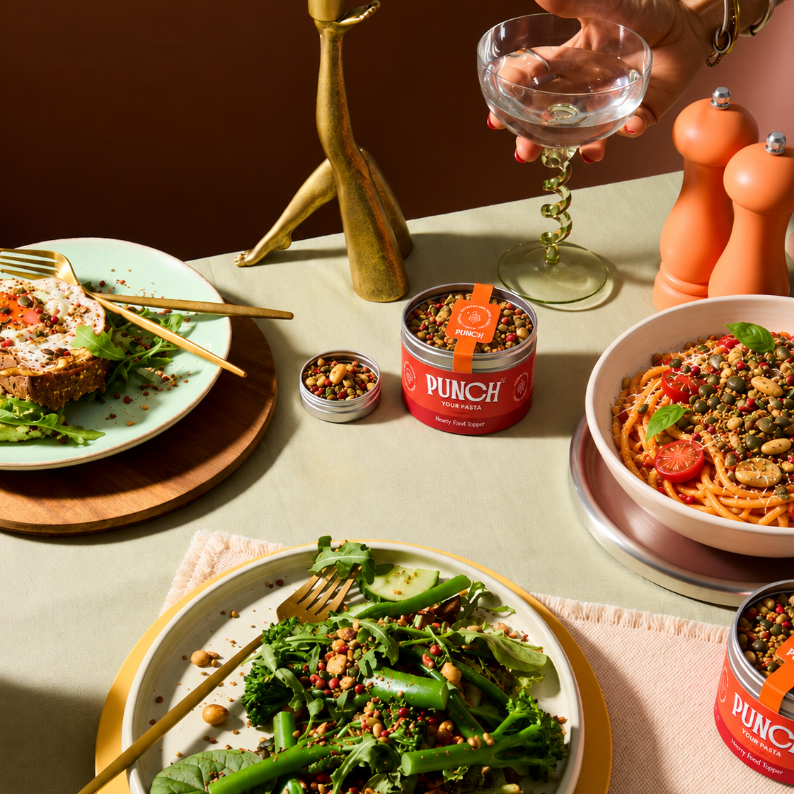 A styled dining table scene featuring PUNCH canned pasta products, with plates of prepared food including pasta, greens, and toast, a hand holding a glass, and warm home dining props against a dark brown background.