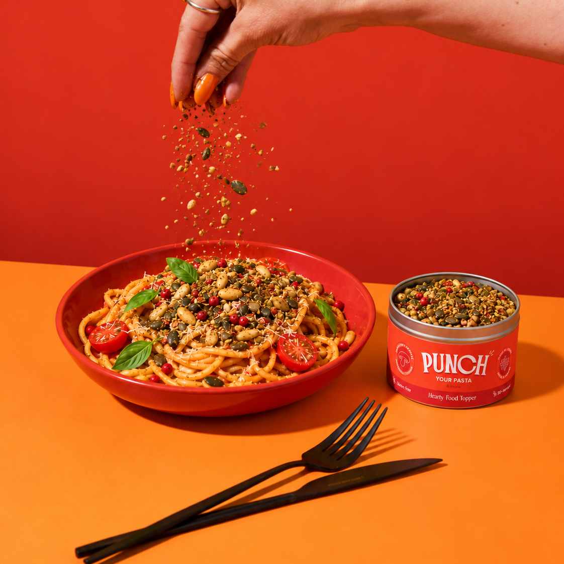 A hand sprinkles dried herbs over a bowl of pasta on an orange table, with a red-orange background and a can of PUNCH Daily Food Sages standing beside the bowl.