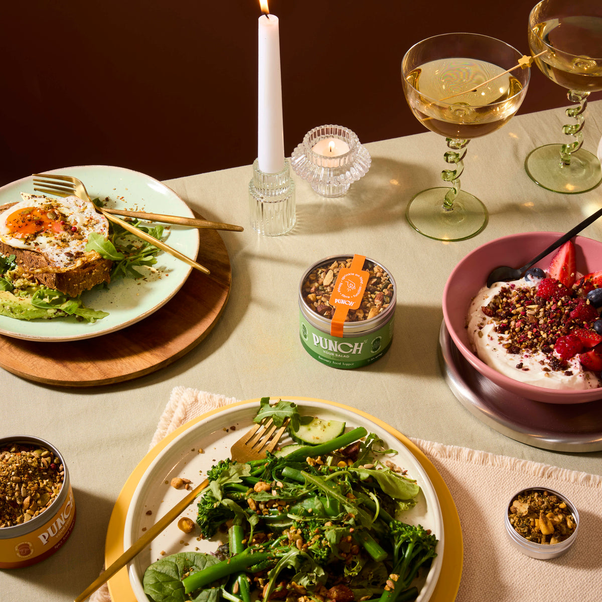 A styled dining table scene with plated food, two stemmed wine glasses, candles, and small jars of granola or seed mix. The central focus is a plate of green broccoli and greens with nuts and seeds, surrounded by other prepared dishes and packaging.