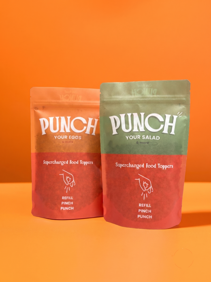 Two stand-up pouches of Punch brand food toppings on a solid orange background. The left pouch is peach and red with white text reading 'PUNCH YOUR EGGS' and 'Supercharged Food Toppers'.