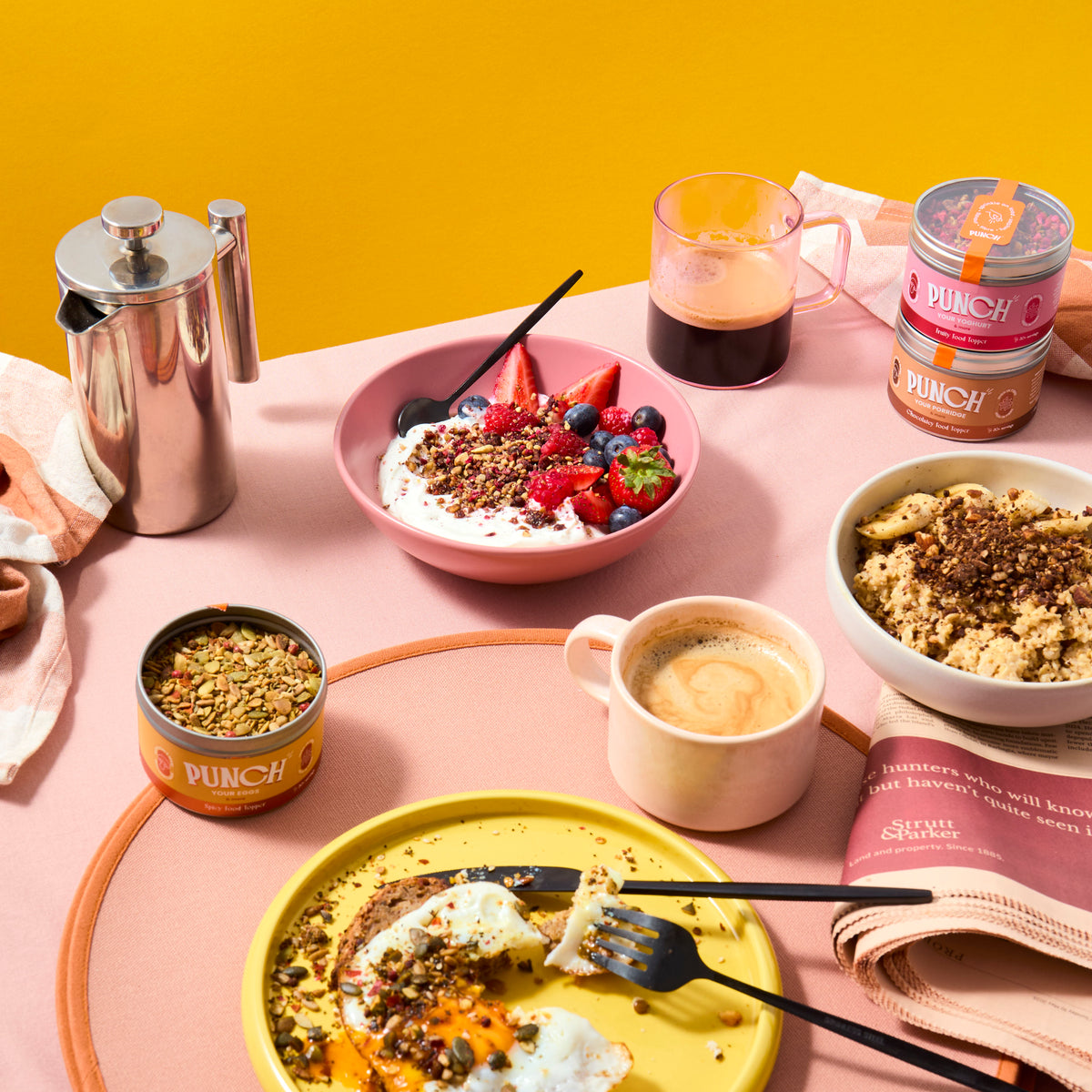 Breakfast table setting with food, coffee, and Punch products on a pink surface with yellow background feeling sweet breakfast scene punch your oats and punch your yoghurt food toppers breakfast scene