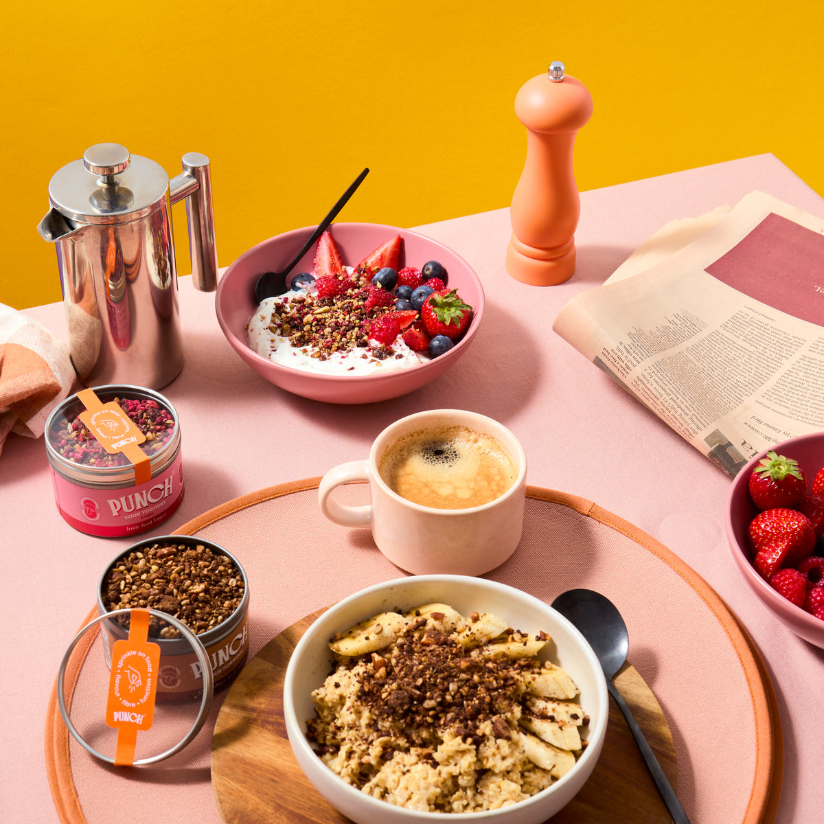 Breakfast scene with punch food toppers punch your oats and punch your yoghurt bowls of food, a coffee cup, and a pink background