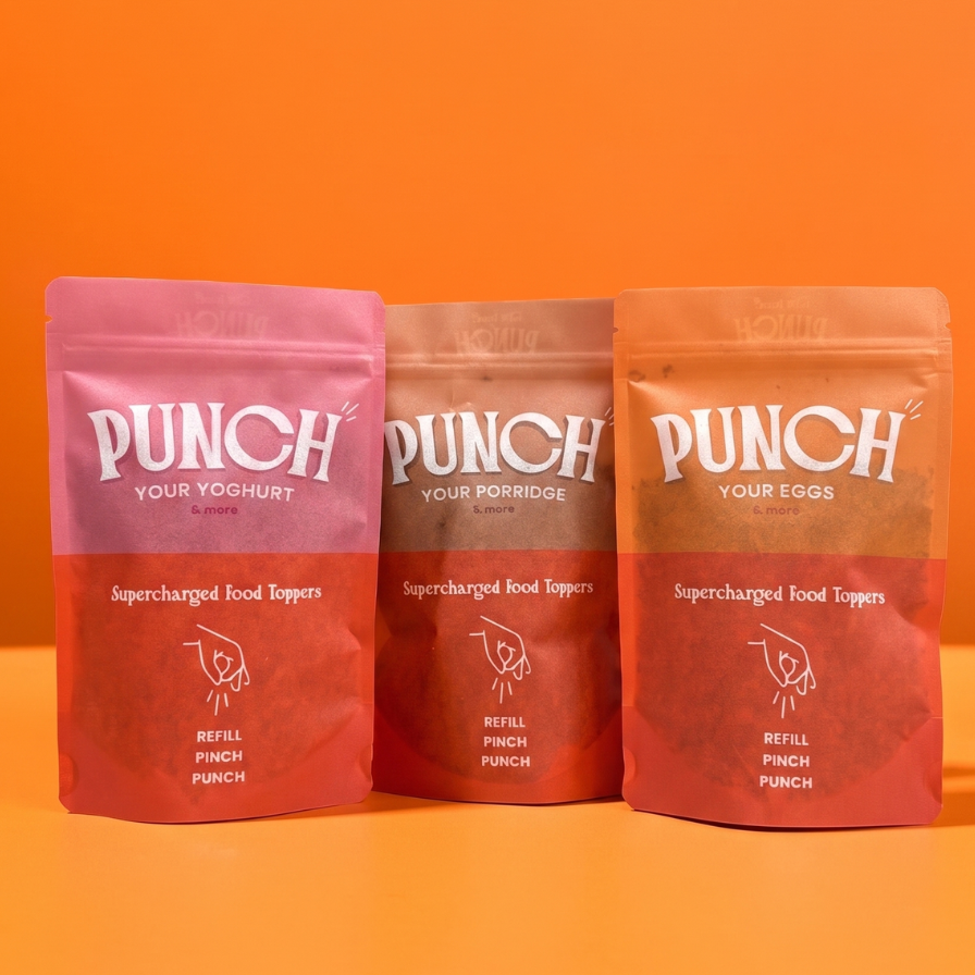 Three stand-up pouches of Punch Your Yogurt / Your Eggs food toppings arranged side by side on an orange surface against an orange background. The left pouch is pink, the middle is brown, and the right is red-orange, all with white branding text.