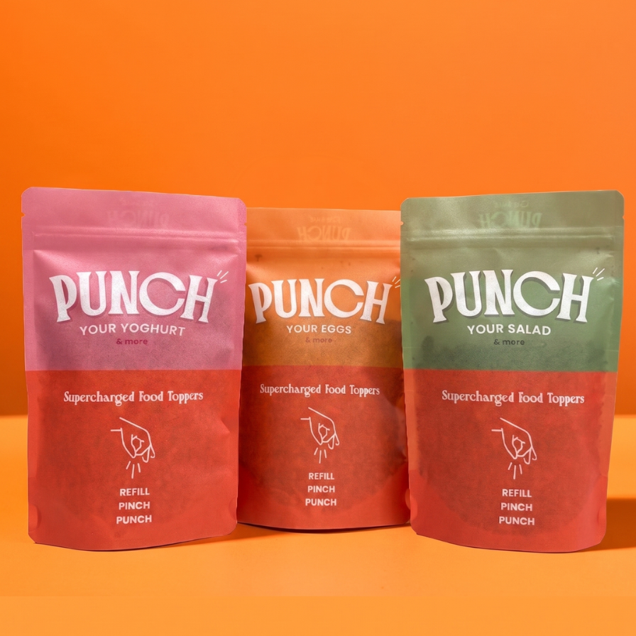 Three stand-up pouches of Punch Your Yogurt/Protein food toppers are arranged on an orange background. The left pouch is pink and labeled 'Punch Your Yogurt', the middle pouch is orange and labeled 'Punch Your Eggs', and the right pouch is green and labeled 'Punch Your Salad'.
