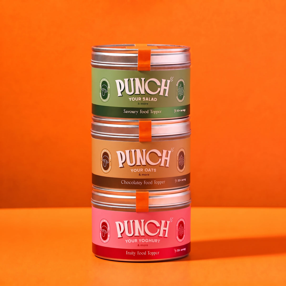 Three stacked tins of PUNCH food toppers on a solid orange background. The top tin is Savory Food Topper in sage green, the middle is Chocolatey Food Topper in tan, and the bottom is Fruity Food Topper in pink.