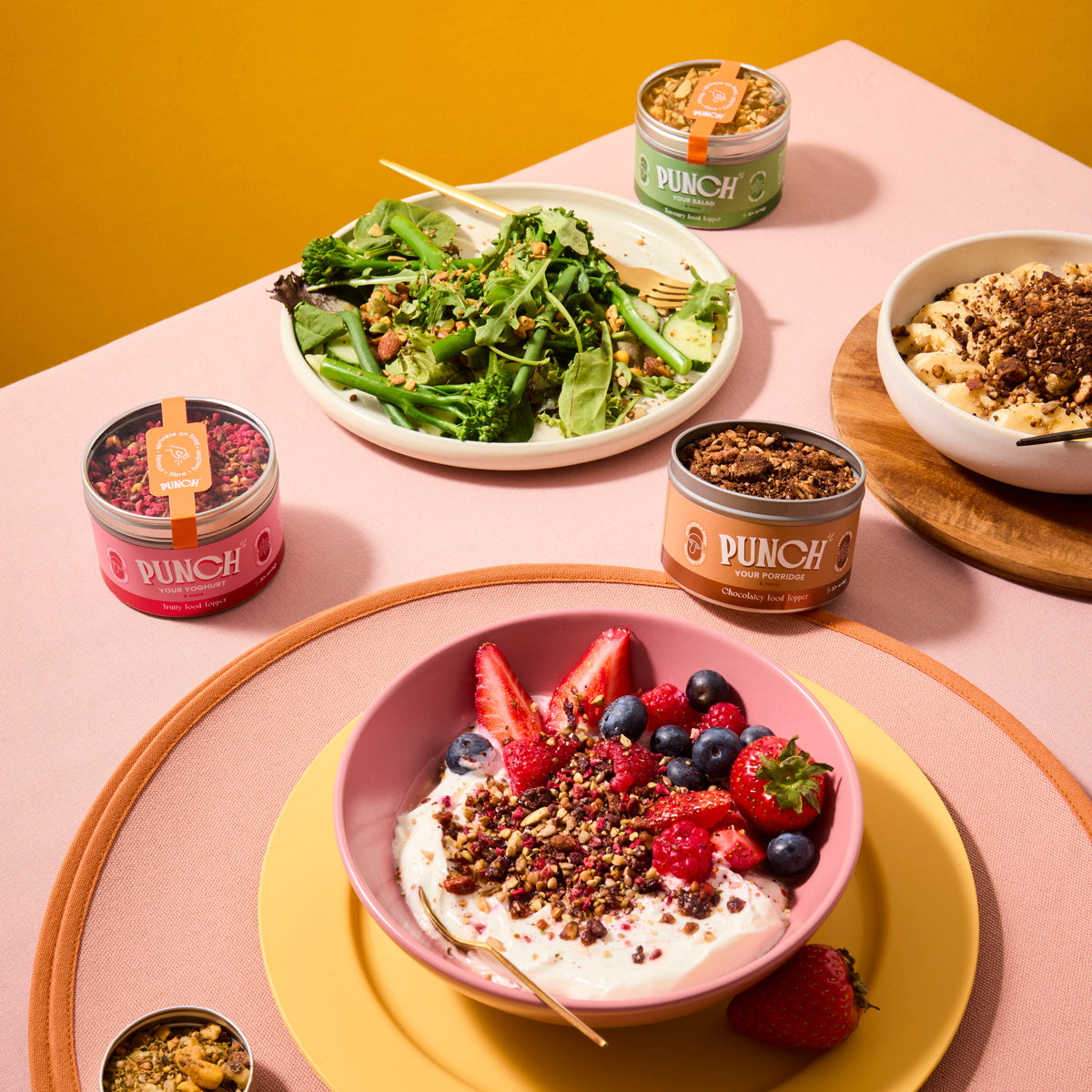Table setting with bowls of salad, yogurt with berries, and Dunch snack tins on a pink surface.