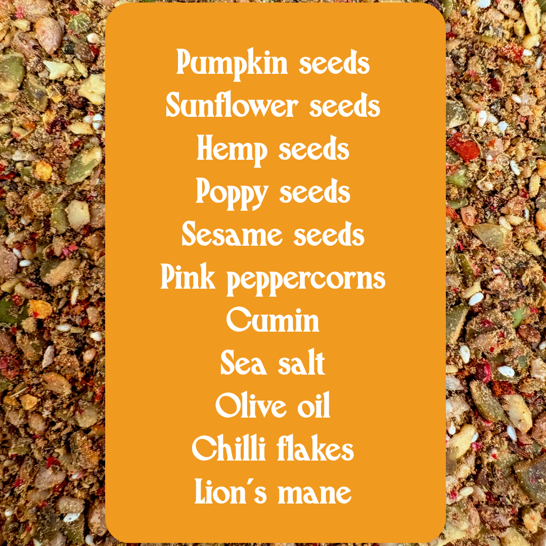 A flat-lay product shot of a spice blend featuring pumpkin seeds, sunflower seeds, hemp seeds, poppy seeds, sesame seeds, pink peppercorns, cumin, sea salt, olive oil, chili flakes, and lion's mane, presented on a textured background with a central orange label listing the ingredients.