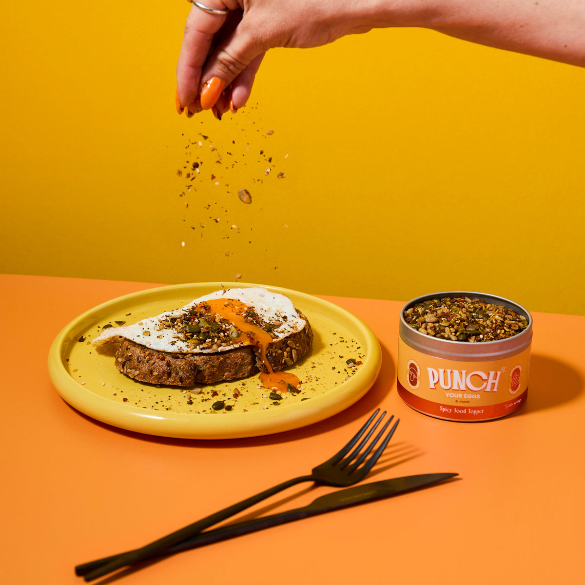 Hand sprinkling seeds over a dish with a can of 'Punch your eggs' spicy food topper on a yellow and orange background