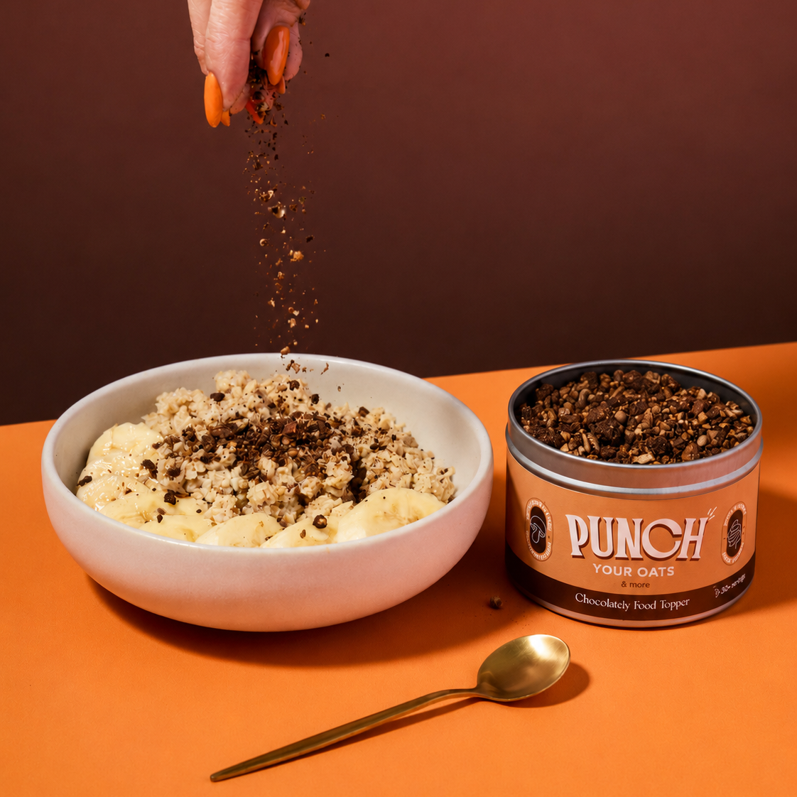 A hand sprinkles chocolate food topper over a bowl of porridge, with a tin of PUNCH Your Oats Chocolate Food Topper and a gold spoon on an orange surface against a dark brown background.