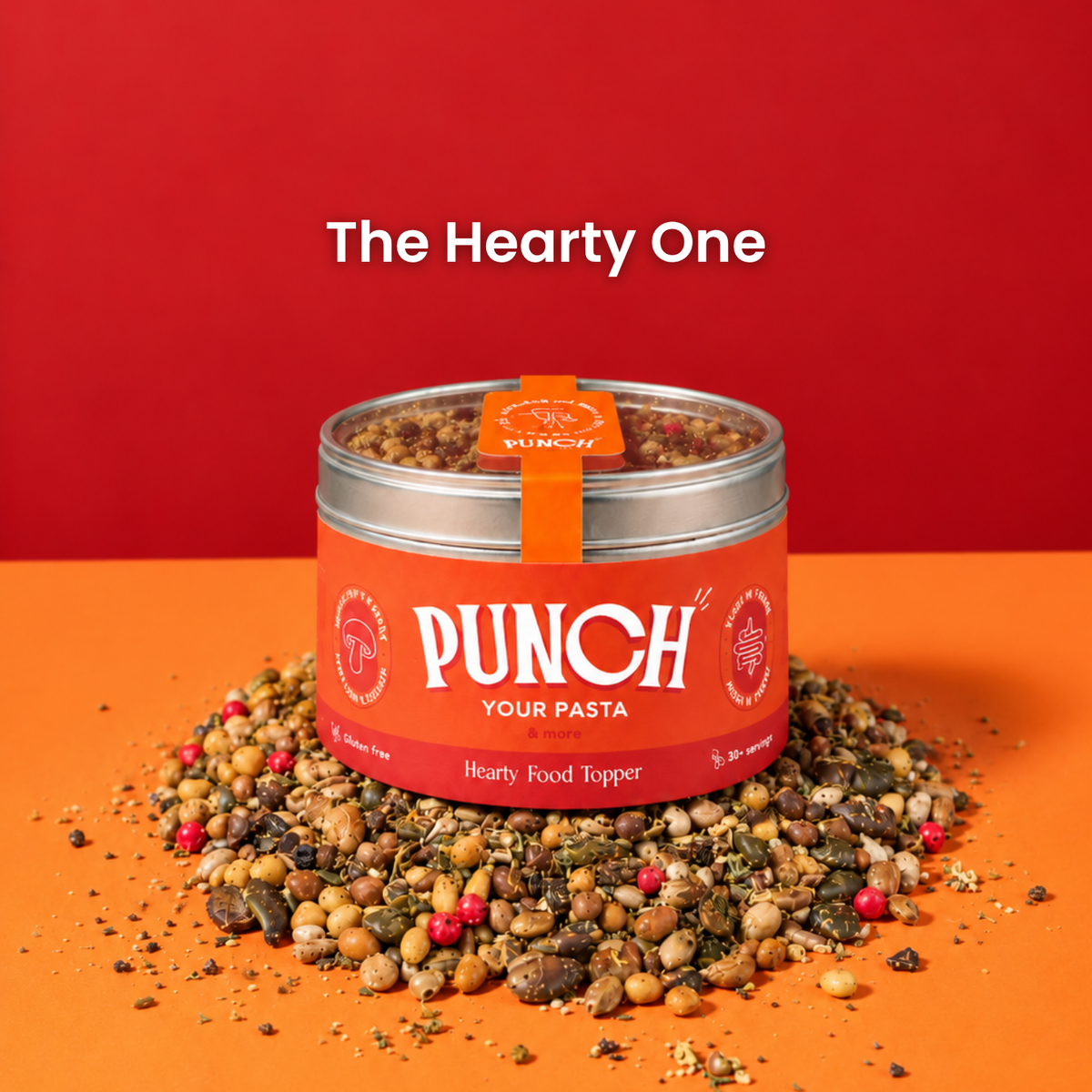 A tin of PUNCH Your Pasta Hearty Food Topper sits on a pile of mixed dried herbs, grains, and spices against a vibrant red and orange background. The text 'The Hearty One' is displayed above the product.
