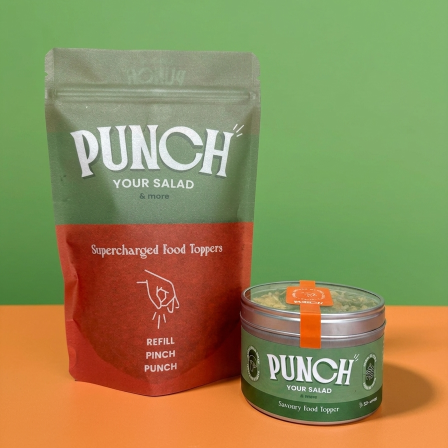 A pouch and a small round tin of PUNCH Your Salad supercharged food toppers on a two-tone green and orange background. The pouch is olive green and red, and the tin is green with a small orange seal.