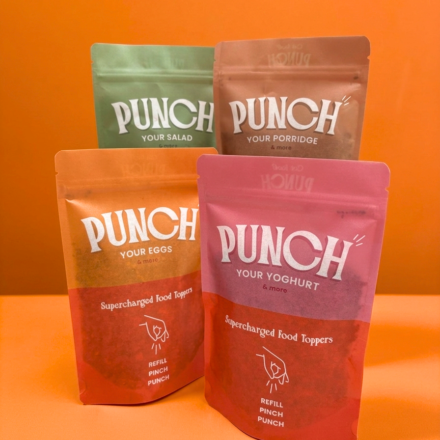 Four stand-up pouches of Punch brand food products arranged on an orange surface against an orange background. The front pouch is pink and labeled Punch Supercharged Food Toppers, with white text.