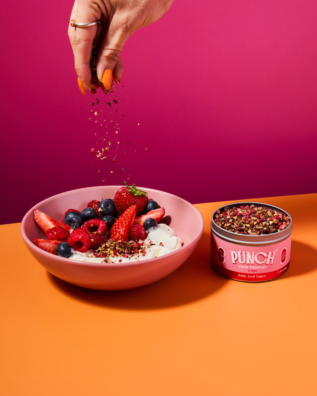 Bowl of fruit salad with yogurt, a tin of Punch your yoghurt fruity food topper, and a hand sprinkling seasoning on a pink and orange background.