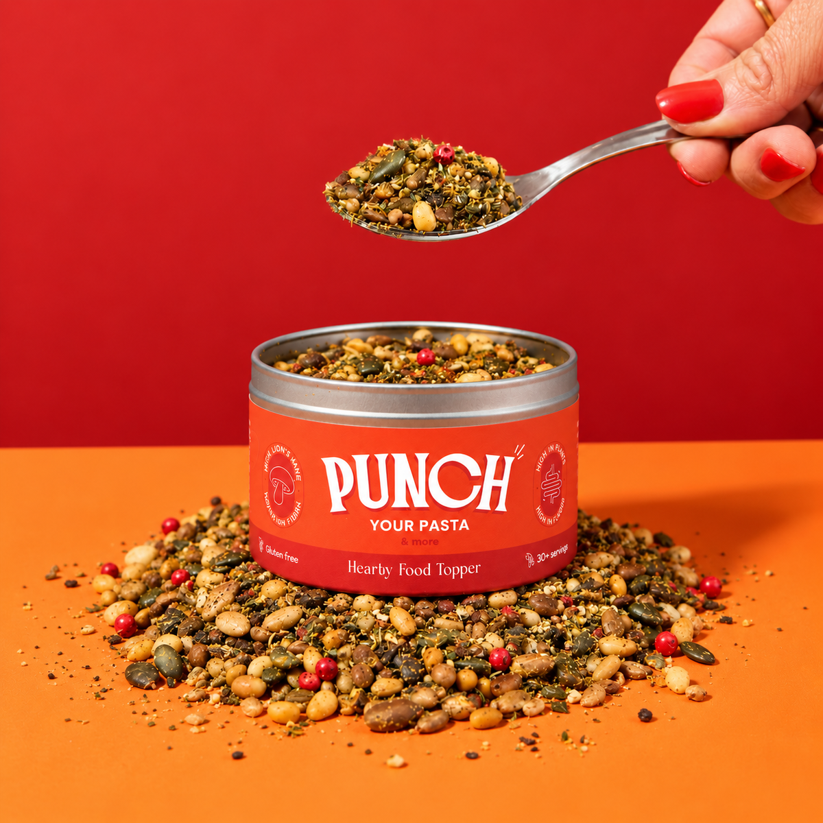 A hand with red nail polish holds a spoon filled with a seed and grain mix above an open red tin labeled PUNCH YOUR PASTA, with loose mix scattered around the base on an orange surface against a red background.