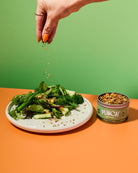 Person adding savoury food topper from a 'Punch your salad' container to a salad on an orange table with a green background