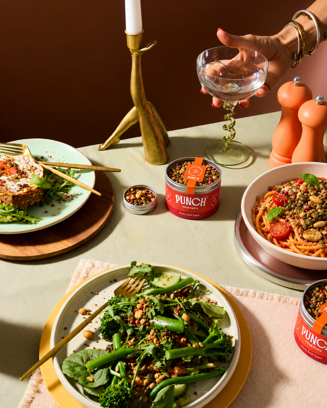 A styled dining table scene featuring PUNCH canned pasta products, with plates of prepared food including pasta, greens, and toast, a hand holding a glass, and warm home dining props against a dark brown background.