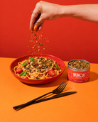 A hand sprinkles dried herbs over a bowl of pasta on an orange table, with a red-orange background and a can of PUNCH Daily Food Sages standing beside the bowl.