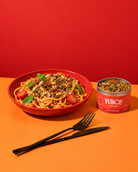 A bowl of pasta topped with herbs, seeds, and cherry tomatoes sits on an orange surface against a red background, next to an open can of DUNCH Posh Pasta. A black fork and knife are placed in front.