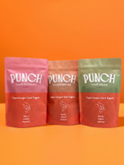 Three stand-up pouches of Punch brand food toppings arranged side by side against a solid orange background. The left pouch is pink, the middle is orange, and the right is green, each with the white 'PUNCH' logo and product text on the front.