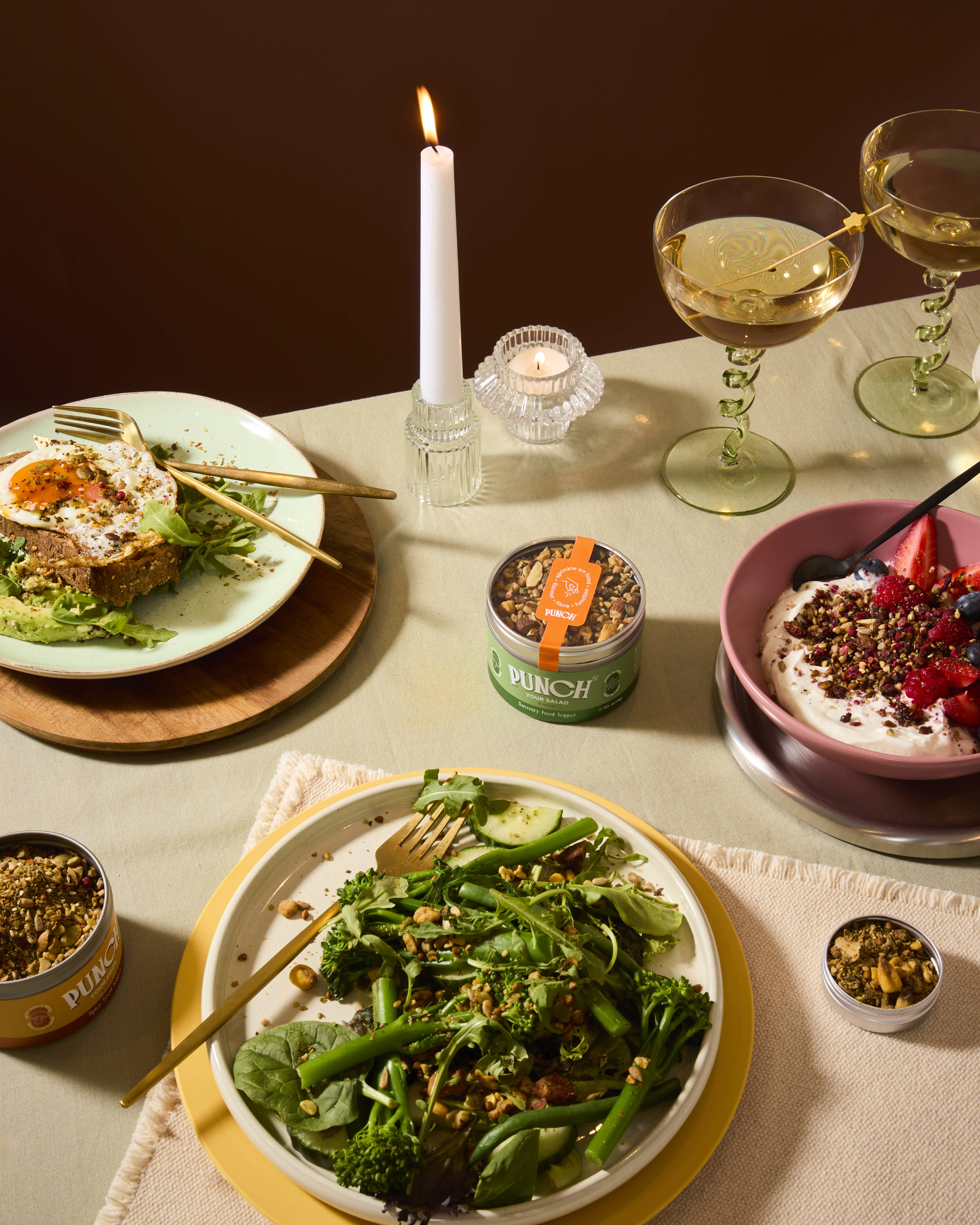 A styled dining table scene with plated food, two stemmed wine glasses, candles, and small jars of granola or seed mix. The central focus is a plate of green broccoli and greens with nuts and seeds, surrounded by other prepared dishes and packaging.