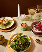 A styled dining table scene with plated food, two stemmed wine glasses, candles, and small jars of granola or seed mix. The central focus is a plate of green broccoli and greens with nuts and seeds, surrounded by other prepared dishes and packaging.