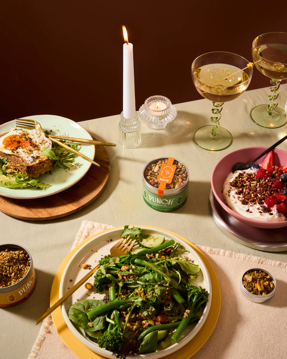 A styled dining table scene with plated food, two stemmed wine glasses, candles, and small jars of granola or seed mix. The central focus is a plate of green broccoli and greens with nuts and seeds, surrounded by other prepared dishes and packaging.