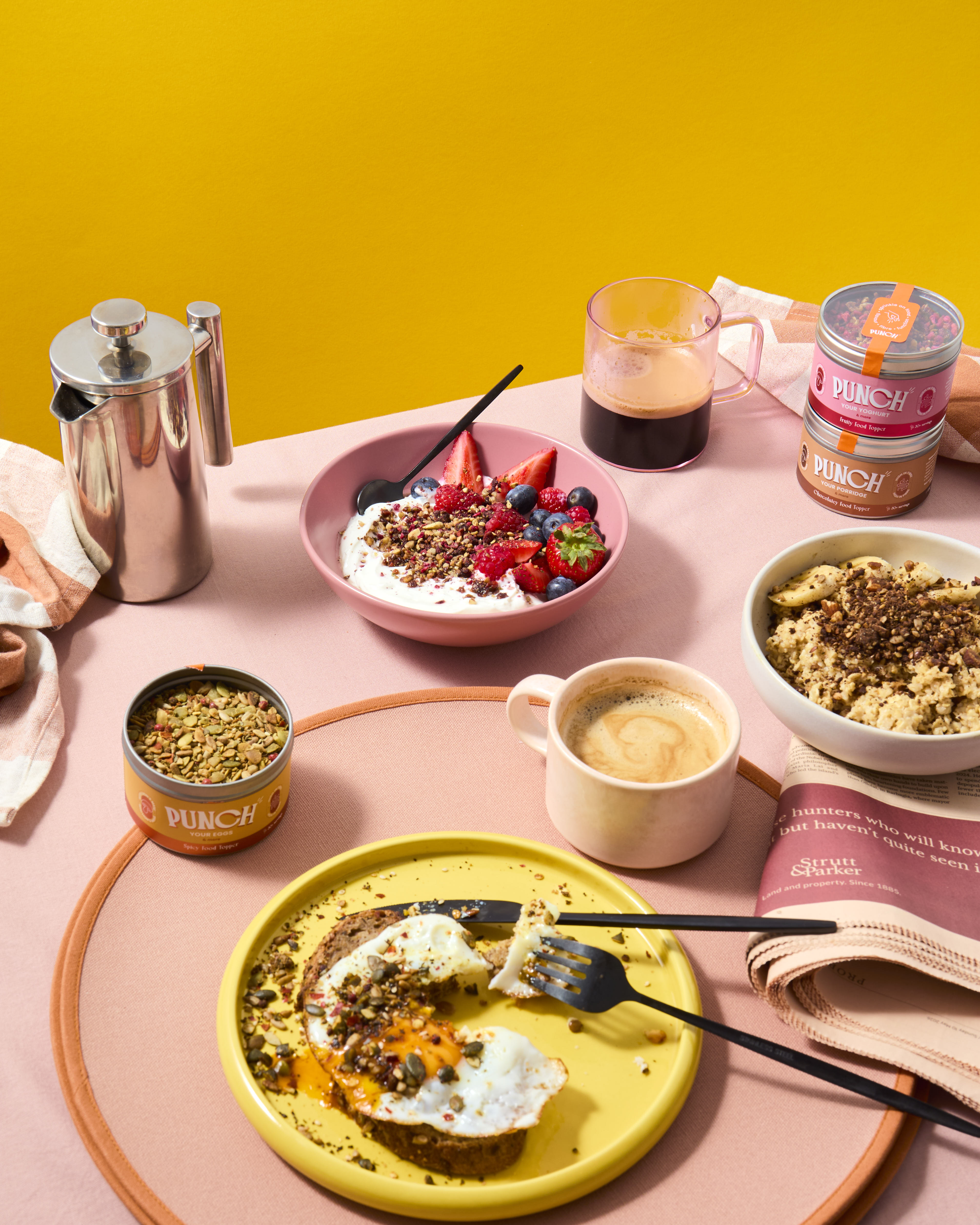 Breakfast table setting with food, coffee, and Punch products on a pink surface with yellow background feeling sweet breakfast scene punch your oats and punch your yoghurt food toppers breakfast scene
