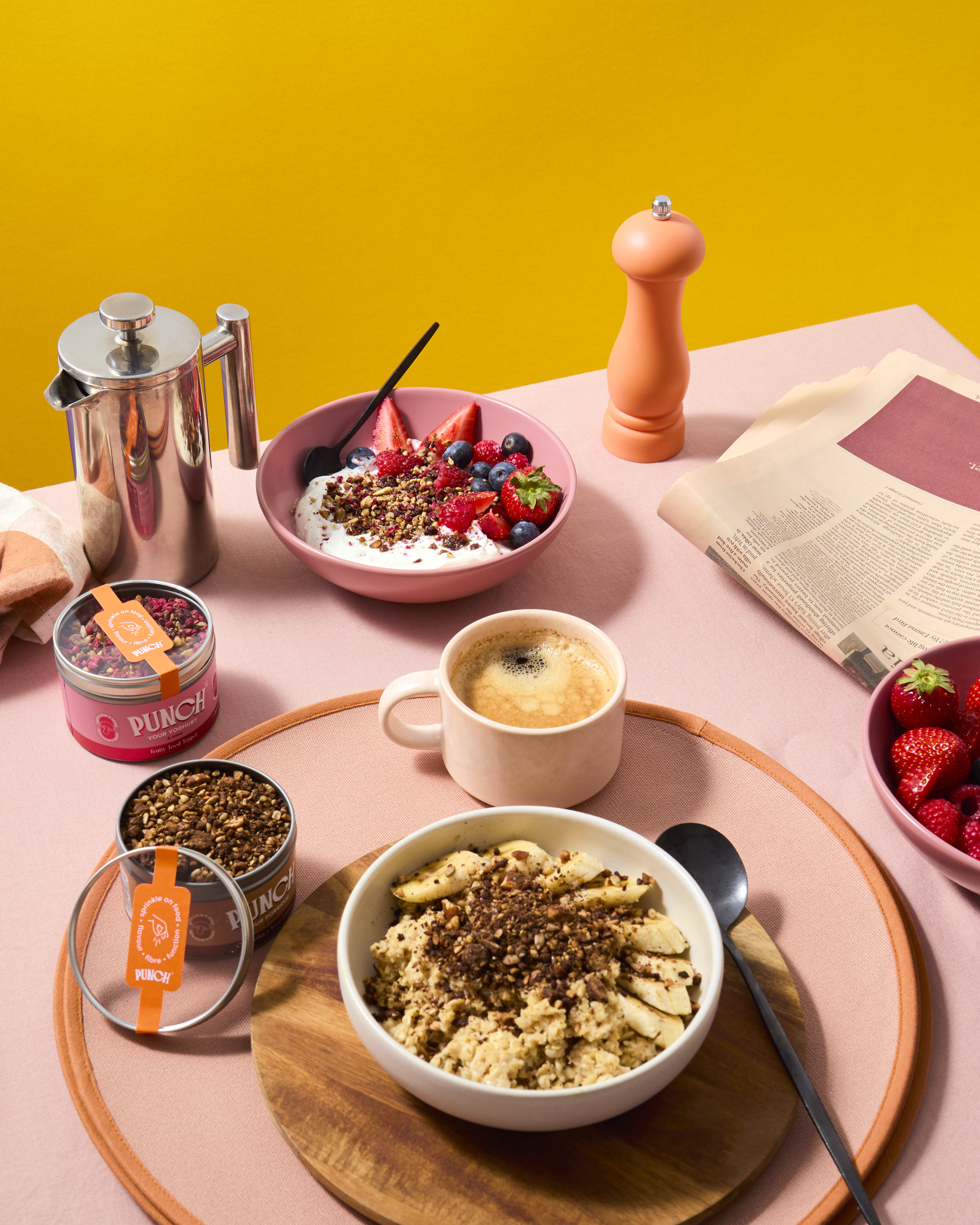 Breakfast scene with punch food toppers punch your oats and punch your yoghurt bowls of food, a coffee cup, and a pink background