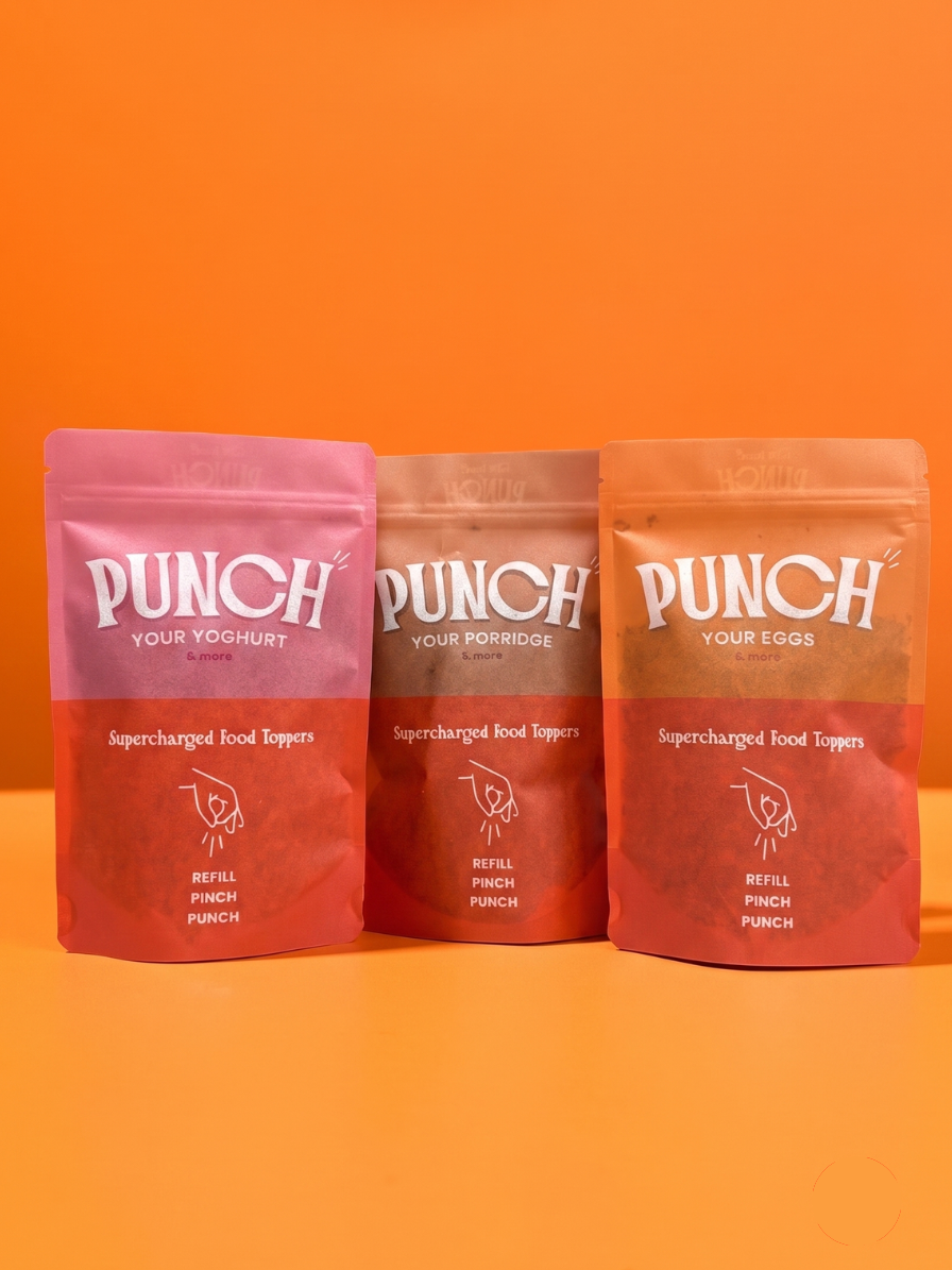 Three stand-up pouches of Punch Your Yogurt / Your Eggs food toppings arranged side by side on an orange surface against an orange background. The left pouch is pink, the middle is brown, and the right is red-orange, all with white branding text.