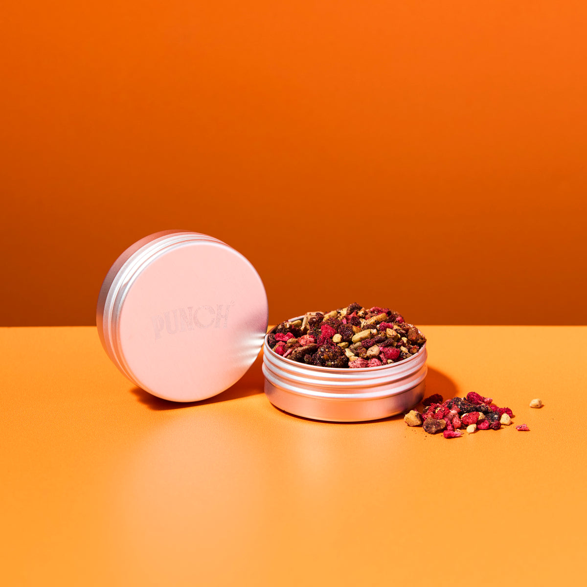 Pink metal tin with a lid open, revealing dried herbs on an orange background