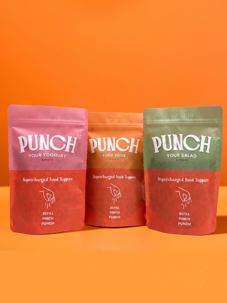 Three stand-up pouches of Punch Your Yogurt/Protein food toppers are arranged on an orange background. The left pouch is pink and labeled 'Punch Your Yogurt', the middle pouch is orange and labeled 'Punch Your Eggs', and the right pouch is green and labeled 'Punch Your Salad'.