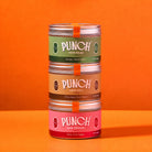 Three stacked tins of PUNCH food toppers on a solid orange background. The top tin is Savory Food Topper in sage green, the middle is Chocolatey Food Topper in tan, and the bottom is Fruity Food Topper in pink.