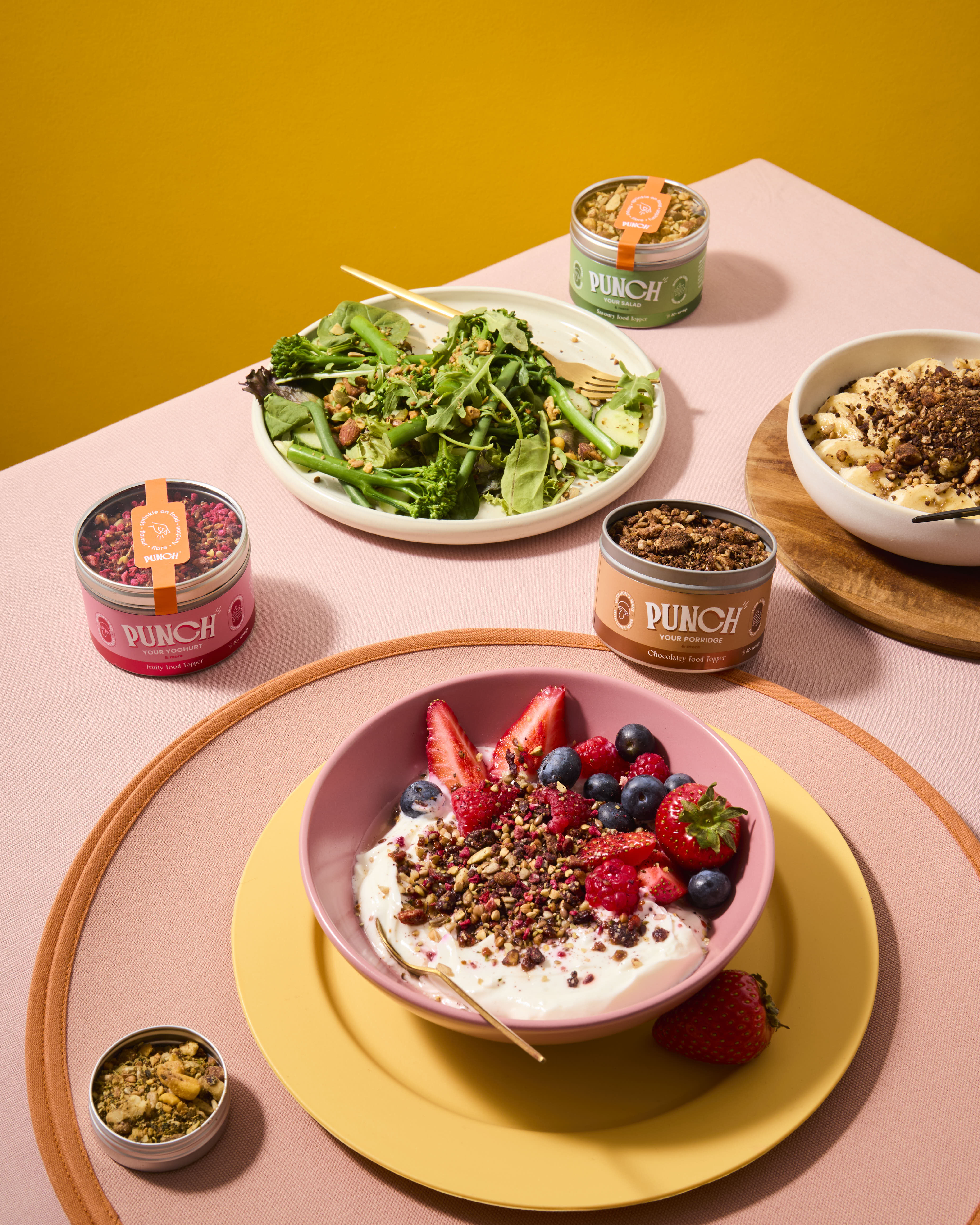 Table setting with bowls of salad, yogurt with berries, and Dunch snack tins on a pink surface.