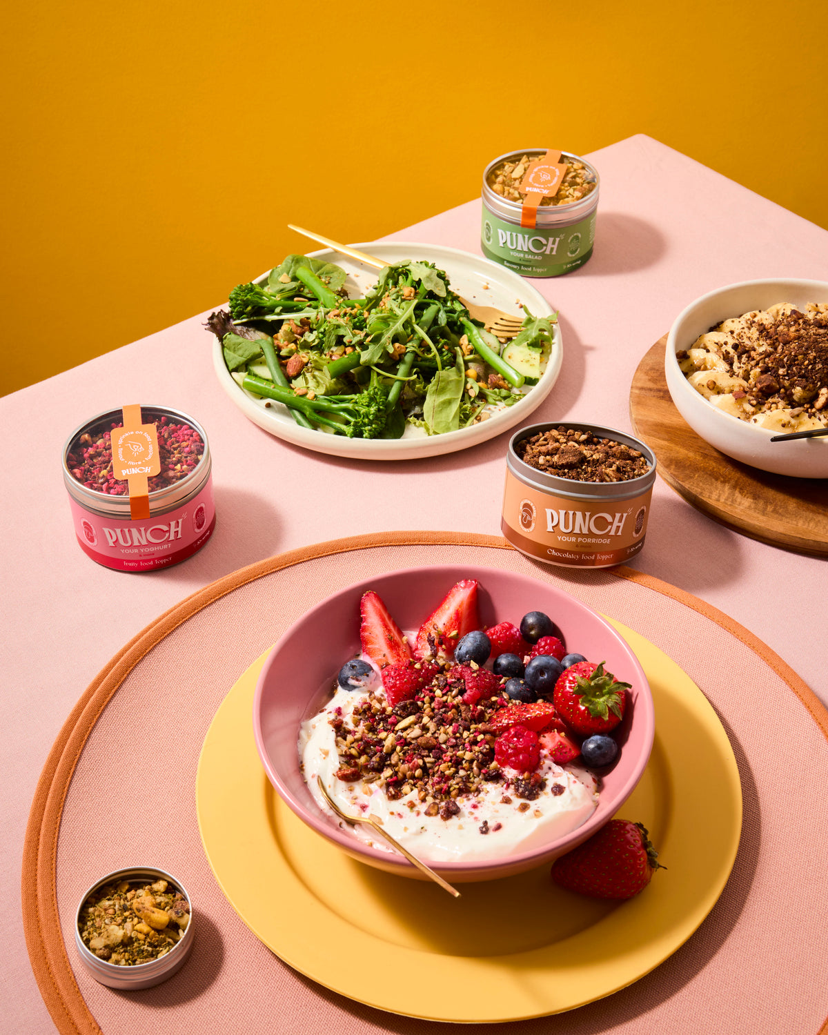 Table setting with bowls of salad, yogurt with berries, and Dunch snack tins on a pink surface.