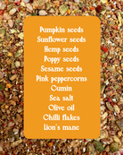 A flat-lay product shot of a spice blend featuring pumpkin seeds, sunflower seeds, hemp seeds, poppy seeds, sesame seeds, pink peppercorns, cumin, sea salt, olive oil, chili flakes, and lion's mane, presented on a textured background with a central orange label listing the ingredients.