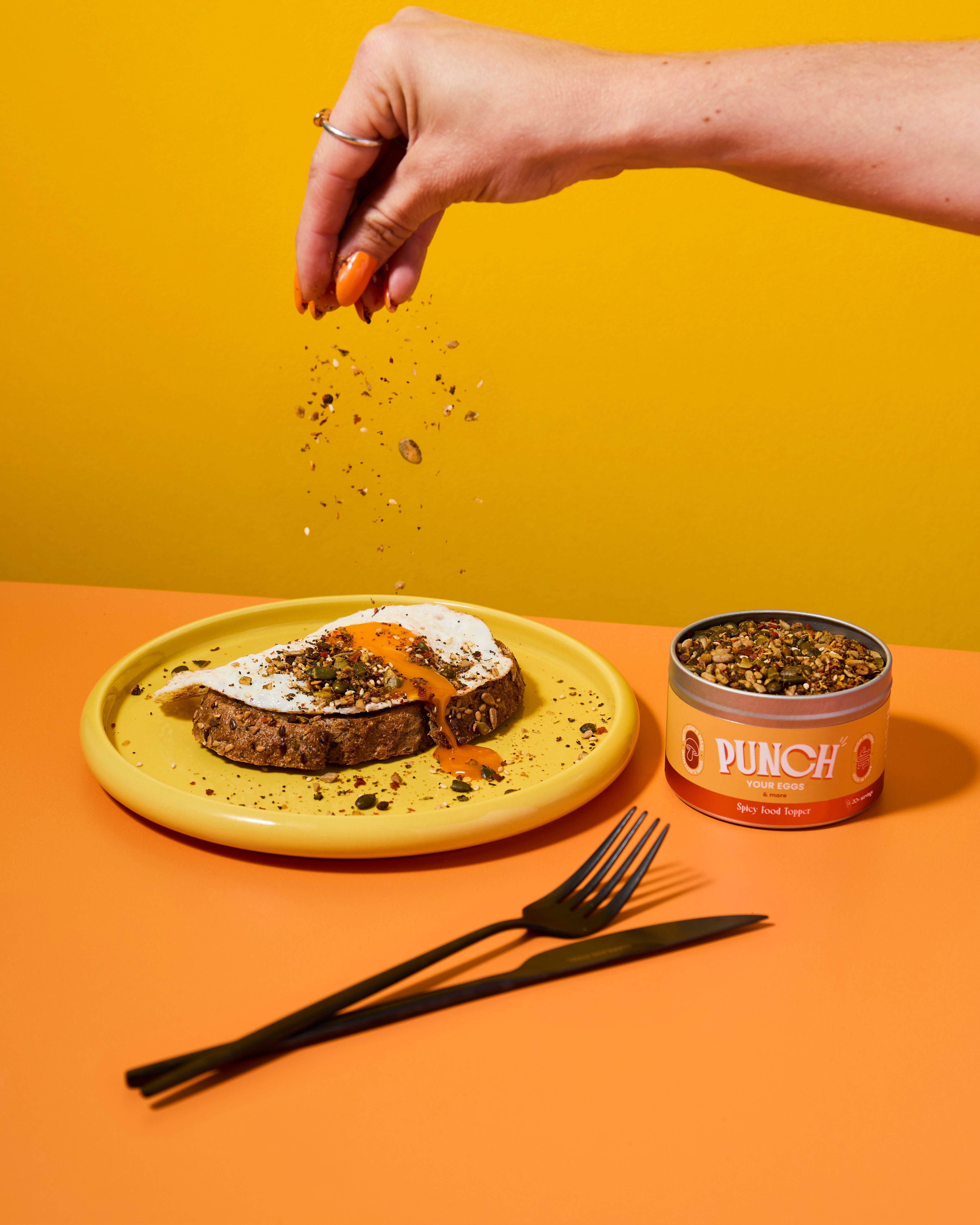 Hand sprinkling seeds over a dish with a can of 'Punch your eggs' spicy food topper on a yellow and orange background