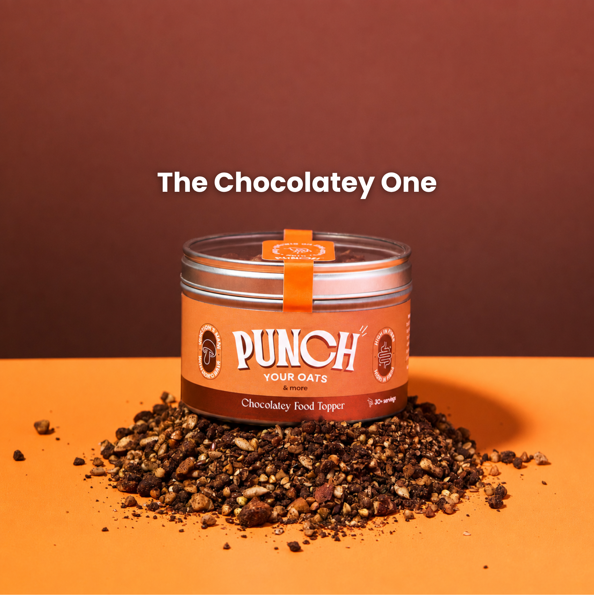 A tin of PUNCH Your Oats Chocolatey Food Topper displayed on an orange surface with a pile of chocolatey oats scattered around it, set against a solid dark brown background with the text 'The Chocolatey One' above.