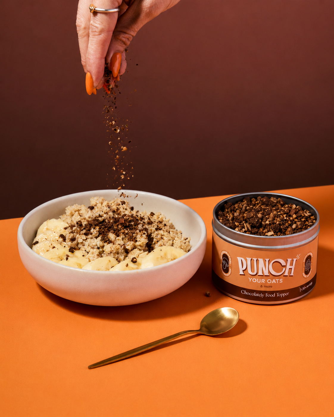 A hand sprinkles chocolate food topper over a bowl of porridge, with a tin of PUNCH Your Oats Chocolate Food Topper and a gold spoon on an orange surface against a dark brown background.