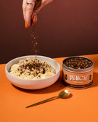 A hand sprinkles chocolate food topper over a bowl of porridge, with a tin of PUNCH Your Oats Chocolate Food Topper and a gold spoon on an orange surface against a dark brown background.