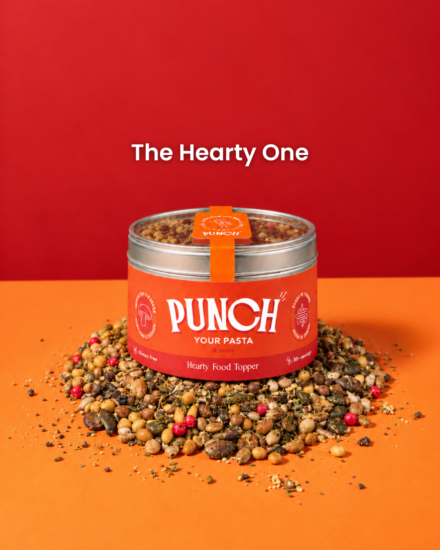 A tin of PUNCH Your Pasta Hearty Food Topper sits on a pile of mixed dried herbs, grains, and spices against a vibrant red and orange background. The text 'The Hearty One' is displayed above the product.