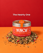 A tin of PUNCH Your Pasta Hearty Food Topper sits on a pile of mixed dried herbs, grains, and spices against a vibrant red and orange background. The text 'The Hearty One' is displayed above the product.