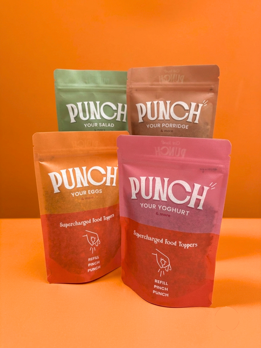 Four stand-up pouches of Punch brand food products arranged on an orange surface against an orange background. The front pouch is pink and labeled Punch Supercharged Food Toppers, with white text.